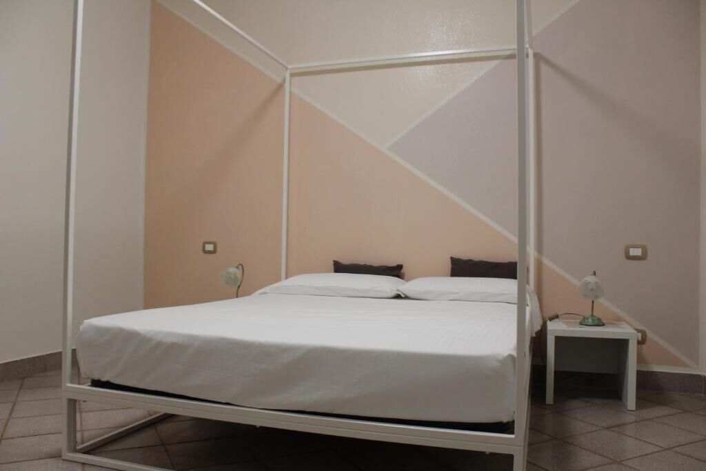 Ein Zimmer mit einem Einzelbett mit weißem Metallgestell, einem kleinen Nachttisch mit Lampe sowie einer Wand mit geometrischem Muster.