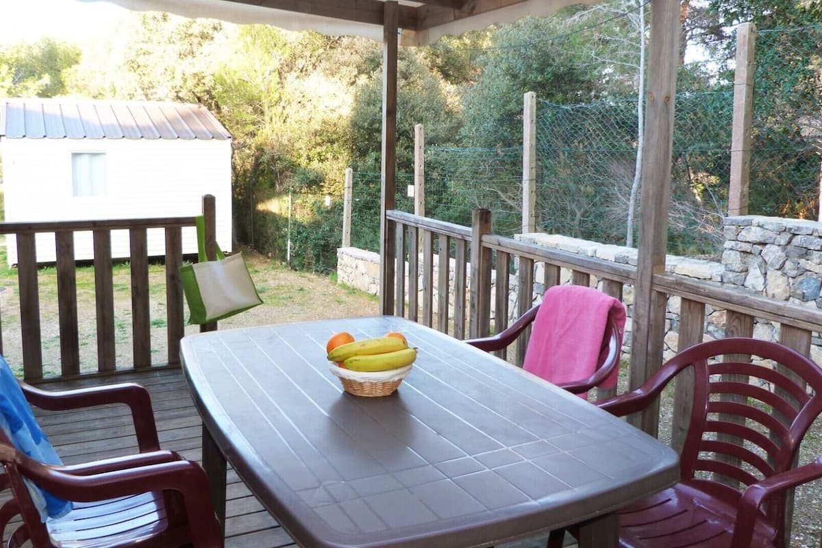 Un porche techado con una mesa y sillas, una cesta de fruta y vistas a una casa y árboles.