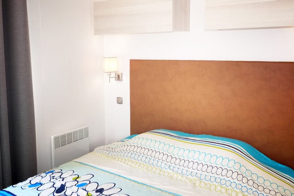 Habitación de hotel con cama, cabecera, luz empotrada en la pared y ventana con cortinas.