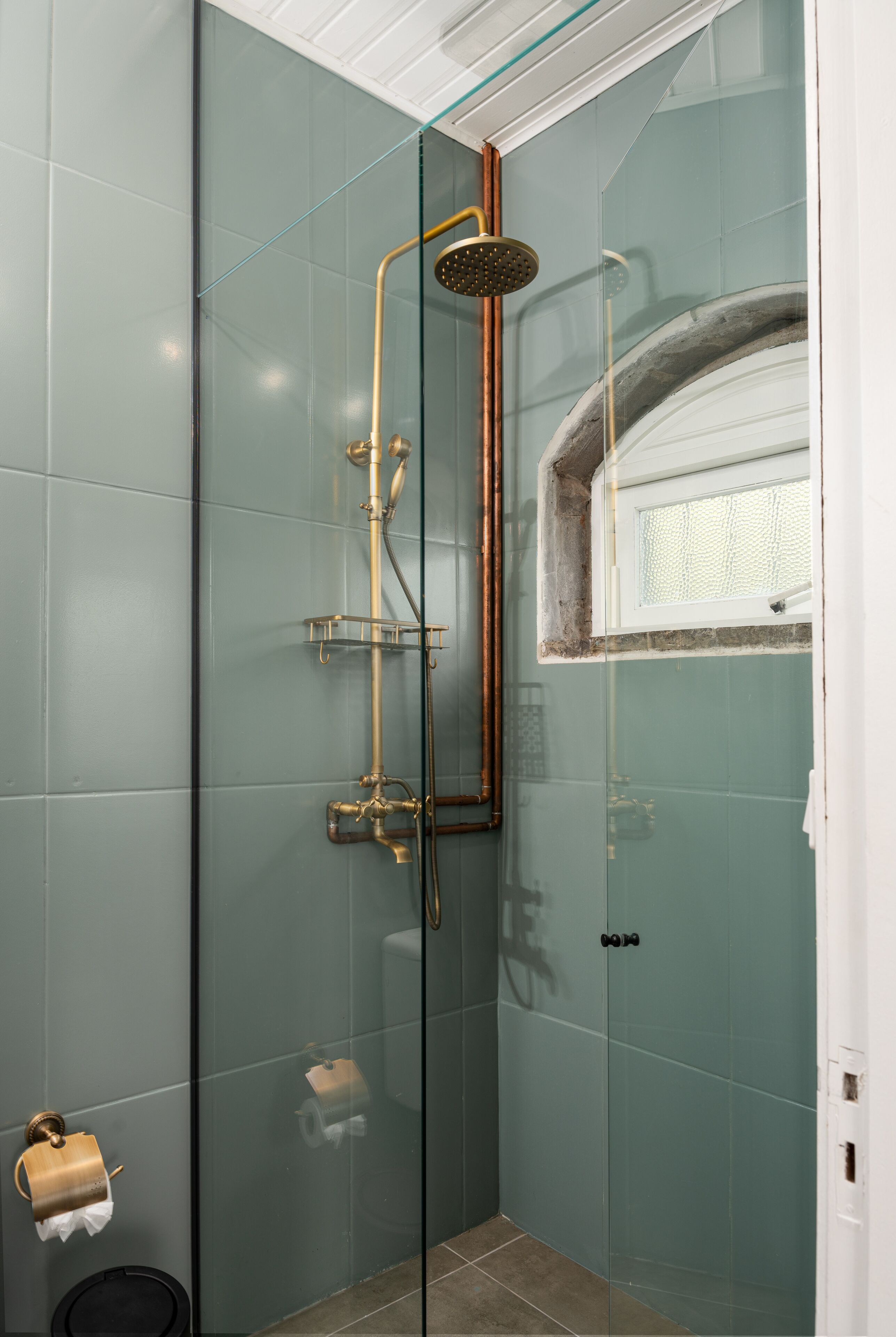 Ducha con cerramiento de vidrio, cabezal y tuberías dorados, un estante pequeño y una ventana.