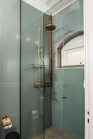 Ducha con cerramiento de vidrio, cabezal y tuberías dorados, un estante pequeño y una ventana.