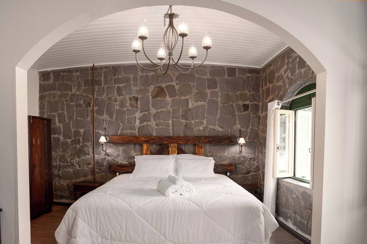 A bedroom with a stone wall, a wooden headboard, a chandelier, and a large bed.