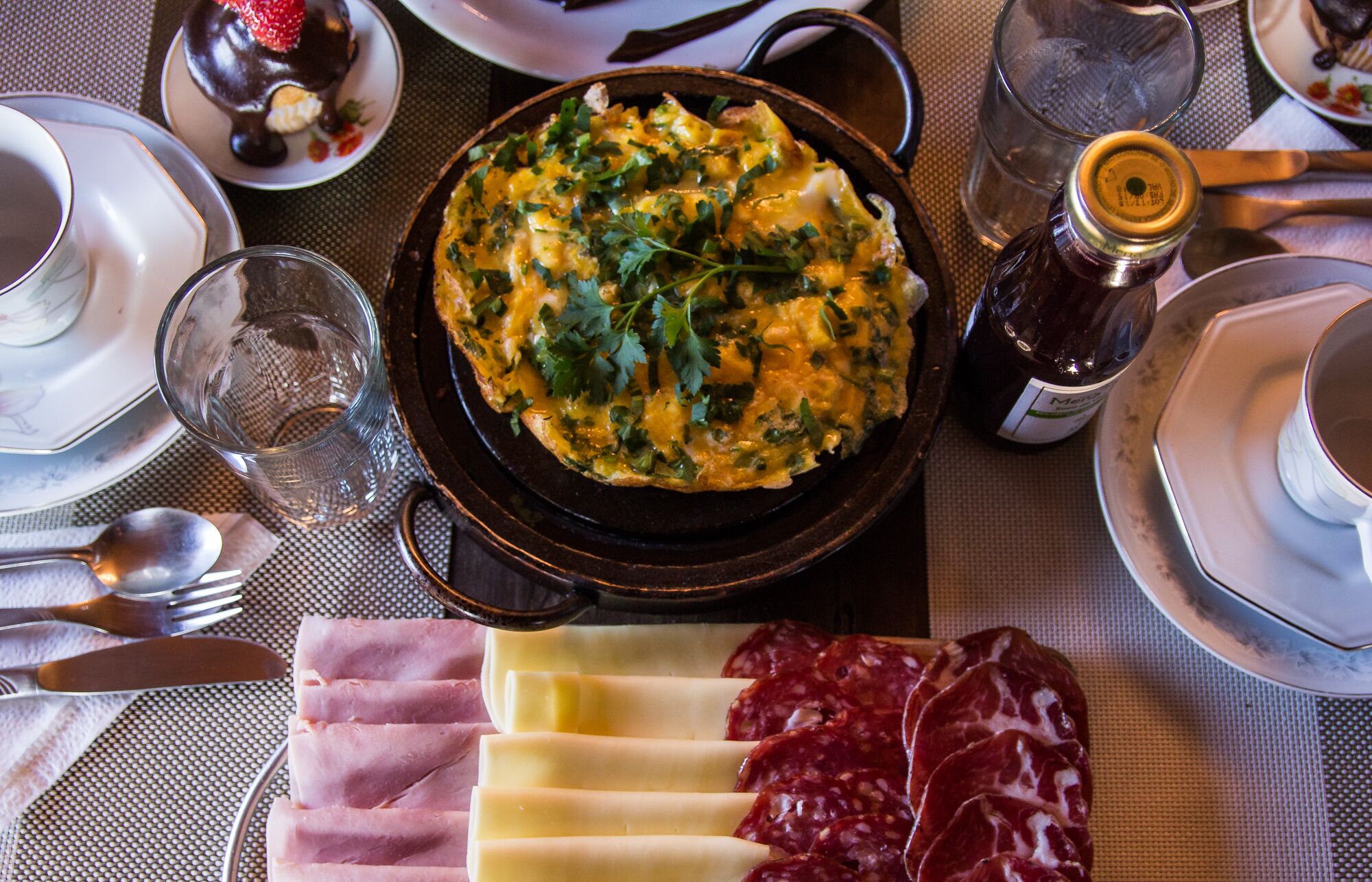 A dining table with a dish of scrambled eggs garnished with herbs, a bottle of wine, and sliced meats.