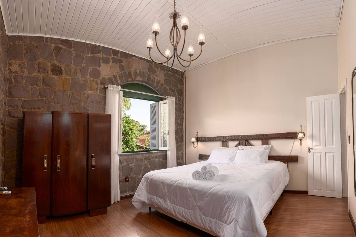 A bedroom with a stone wall, wooden wardrobe, a bed with white bedding, and a chandelier.