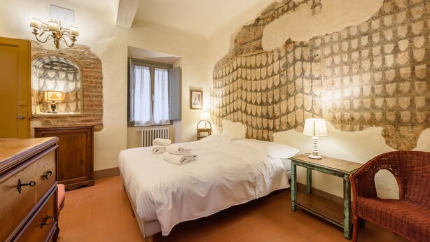 A bedroom with a large bed, a dresser, a chair, and a stone wall. - Casa torre via de' Neri by Mmega (Florence)