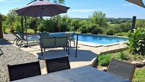 Un espace aménagé au bord de la piscine, avec une table et des chaises, une pergola dotée d’un parasol, et une vue sur la piscine et la verdure environnante.