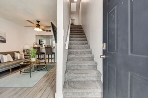 Een trap naar een tweede verdieping, een donkere deur, een woonkamer met een bank, een salontafel en een plafondventilator.