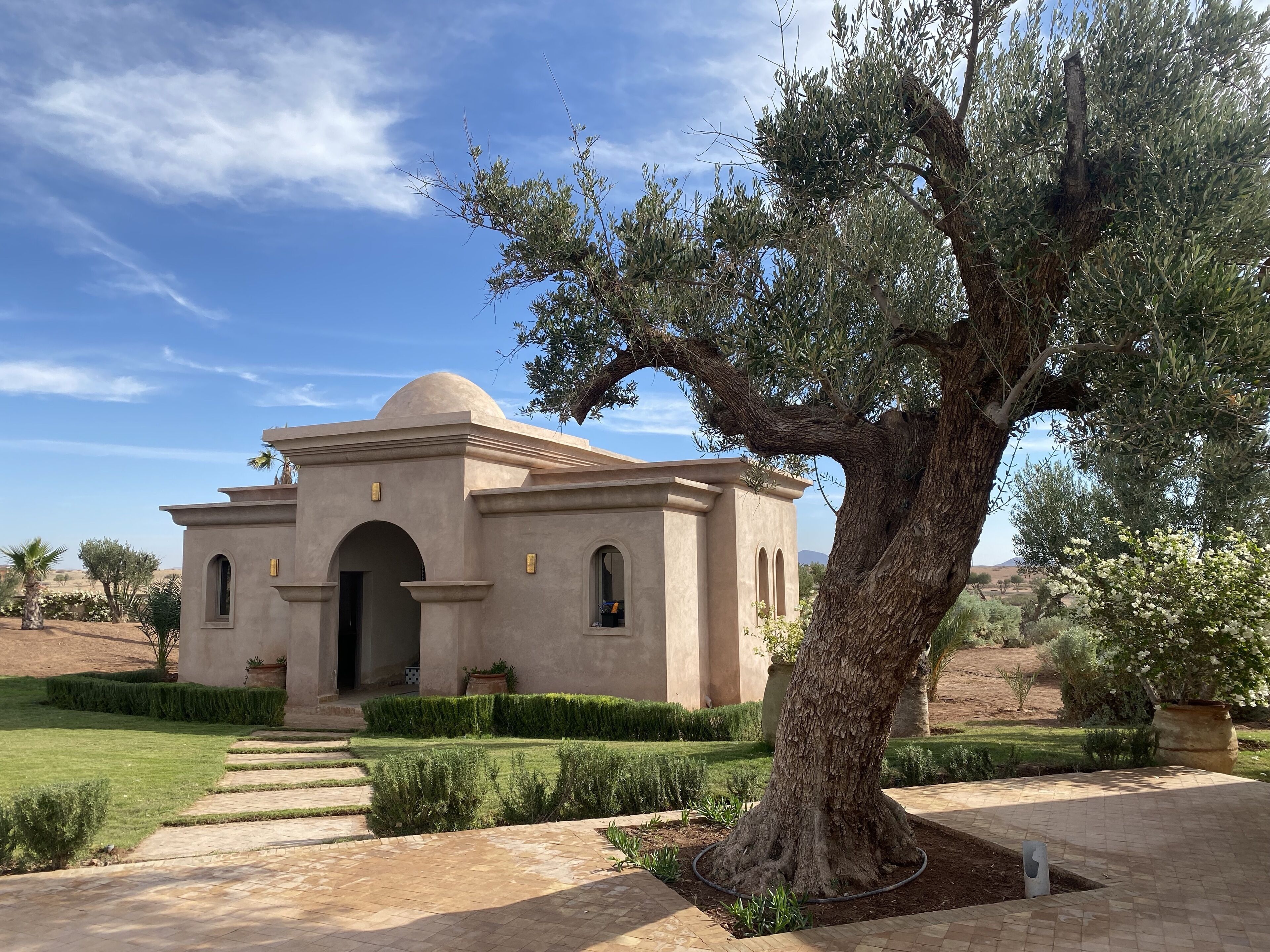 Un bâtiment doté d’un dôme et d’une entrée en arc de cercle, entouré d’un jardin bien entretenu et d’un grand arbre.