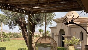 A hanging chair under a wooden pergola, a stone building with arches, and a well-maintained garden with a palm tree and a large pot.
