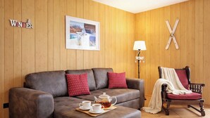 Ein gemütliches Wohnzimmer mit einem braunen Ledersofa, einem Holztisch mit Teekanne und Tassen, einem Holzstuhl mit rotem Kissen, einem gerahmten Bild an der Wand und einem Holzboden.