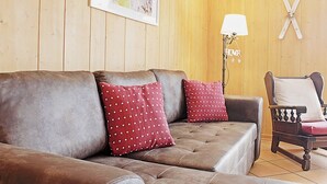 Ein gemütliches Zimmer mit einer braunen Ledersofa, zwei roten Kissen mit weißen Herzen, einem Holzstuhl mit weißem Sitzkissen, einem gerahmten Foto an der Wand und einer Lampe mit der Aufschrift 'HOME'.
