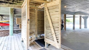 A covered wooden structure with a slatted roof, possibly a sauna or a similar facility, with a concrete floor and a wooden deck in the foreground.