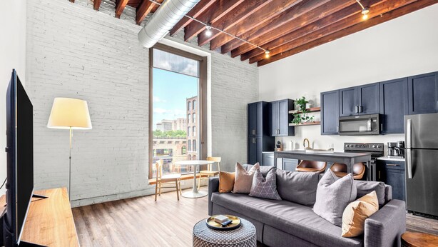 A modern living room with a grey sofa, a coffee table, and a TV. There is a kitchen area with dark cabinets and a stainless steel refrigerator. - Riverfront Loft - Arch Views - Onsite Fitness Center (St. Louis)