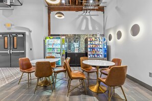 Un espace repas moderne avec une table ronde et six chaises marron, un réfrigérateur rempli d’en-cas et un distributeur automatique.