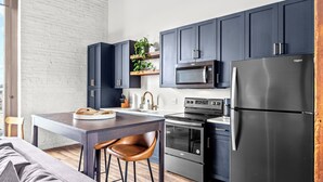 A modern kitchen with dark cabinets, a stainless steel refrigerator, a microwave, and an oven. There is a wooden table with chairs and a plant on the counter. - Riverfront Loft - Arch Views - Onsite Fitness Center (St. Louis)