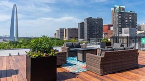 A rooftop terrace with wicker furniture, a wooden deck, and a city skyline view. - Riverfront Loft - Arch Views - Onsite Fitness Center (St. Louis)