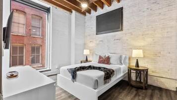 A modern bedroom with a white bed, two bedside tables with lamps, a large window, and exposed wooden beams on the ceiling.