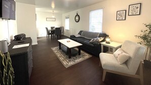 Un salon doté d’un parquet brun foncé, d’une table basse blanche, d’un canapé noir, d’un fauteuil à rayures et d’une petite table d’appoint avec une lampe.