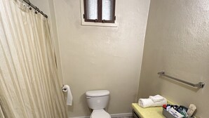 A bathroom with a toilet, a small window, a shower curtain, a towel rack, a sink, and a storage basket. - Cozy Bach Suite  (Winifred)