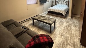 A compact hotel room with a bed, a sofa, a small table, a TV mounted on the wall, and a ceiling light. - Cozy Bach Suite  (Winifred)