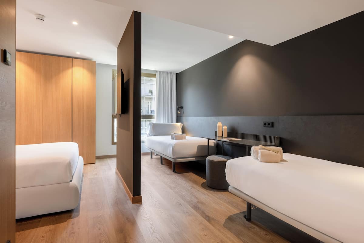 A modern hotel room with a wooden wardrobe, two beds, a bench, a small table with toiletries, and a view of the city.