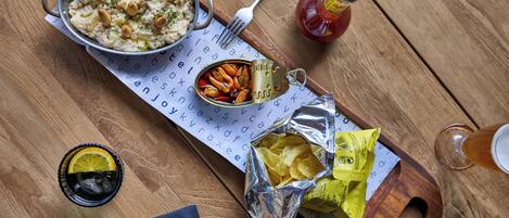Een houten tafel met een schaal eten, een blik sardientjes, een fles ketchup, een glas donker drankje en een servet.