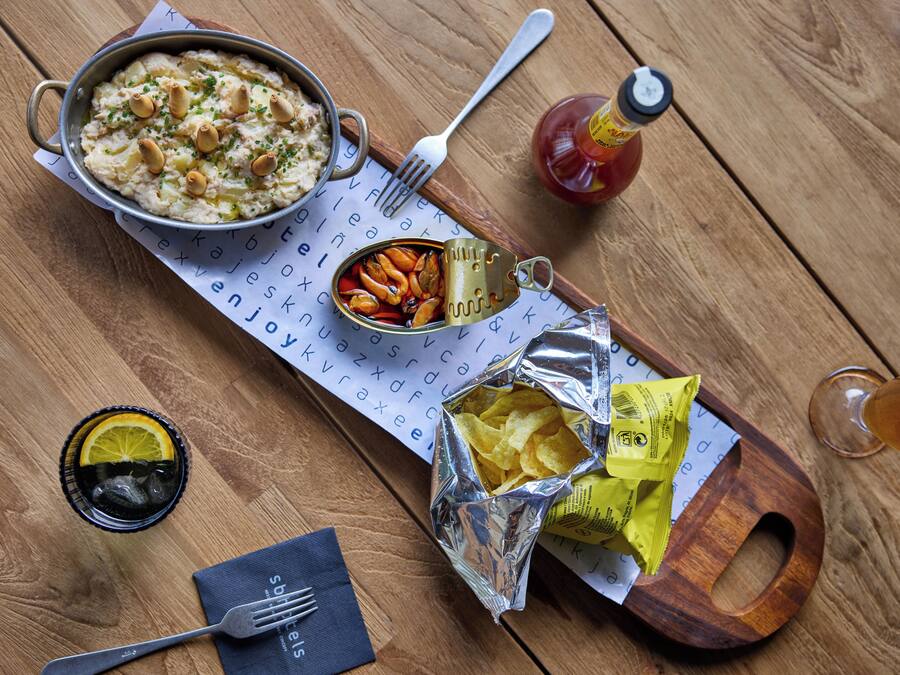 Una mesa de madera con un tazón de comida, una lata de sardinas, una botella de kétchup, un vaso de bebida oscura y una servilleta.