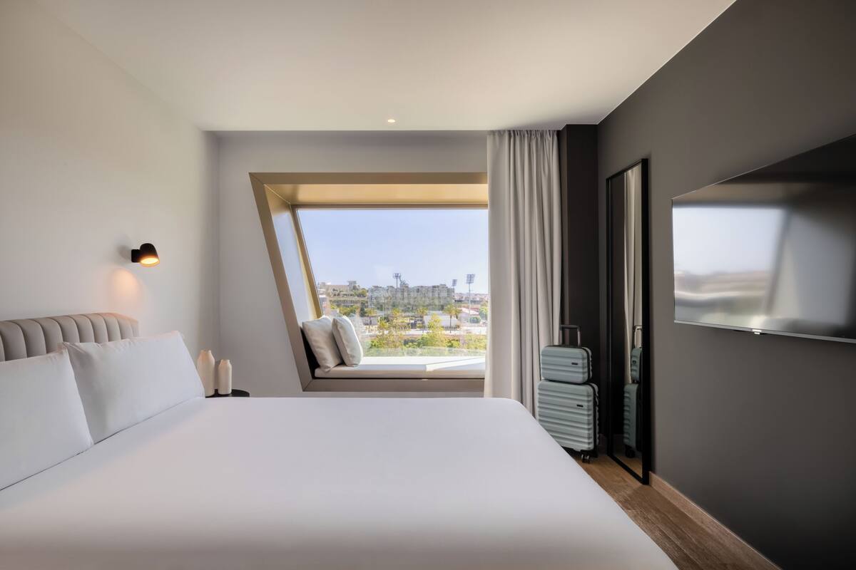 A modern hotel room with a large bed, a TV, a view of the cityscape, and a window with curtains.
