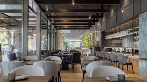 Un restaurante moderno con mesas de madera, sillas grises y una zona de bar con cristalería.