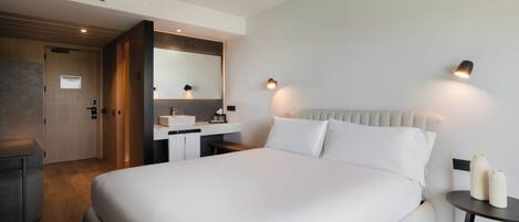 A modern hotel room with a large bed, a bedside table, a mirror, a desk with a chair, and a small round table with two vases.