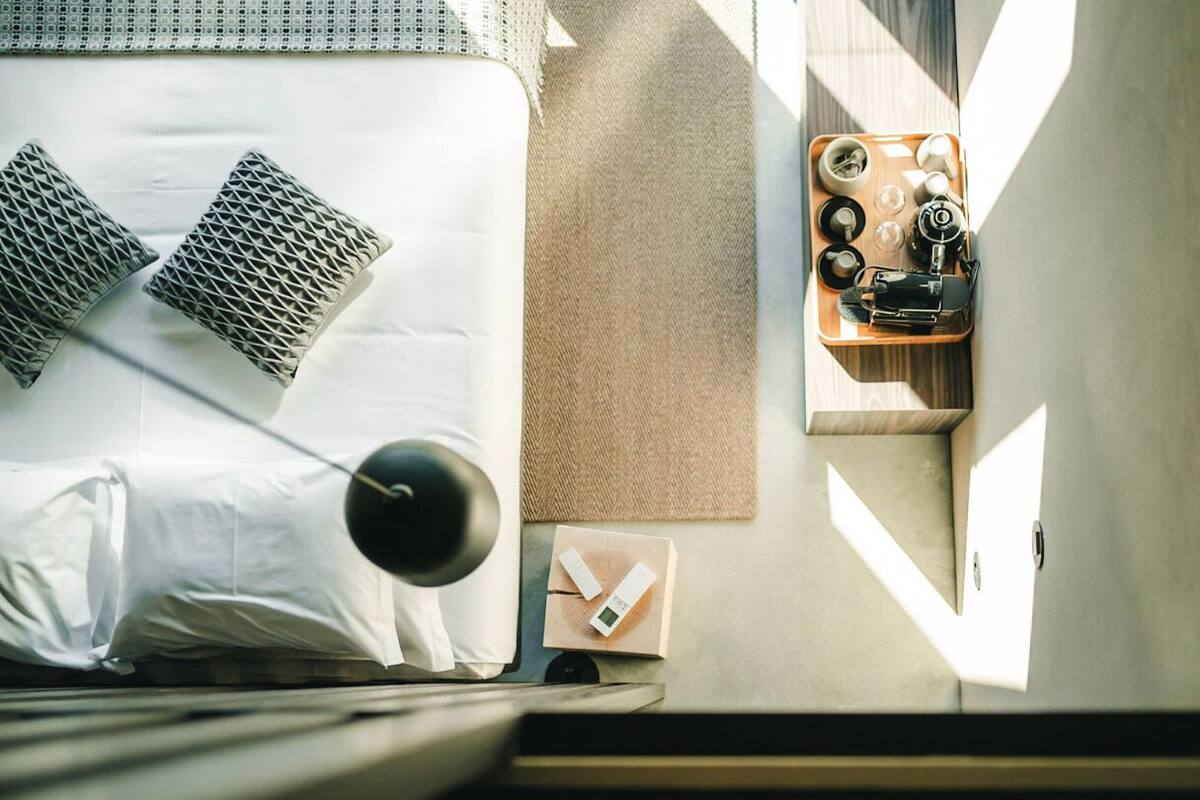 Un lit bien fait, avec des oreillers, une table de chevet avec une lampe, un plateau avec des tasses et un appareil photo, et un luminaire fixé au mur.
