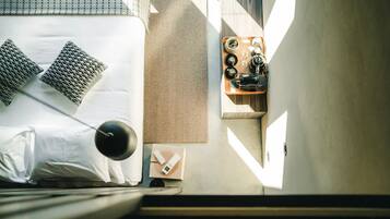 Een net opgemaakt bed met kussens, een nachtkastje met een lamp, een dienblad met kopjes en een camera, en een wandlamp.