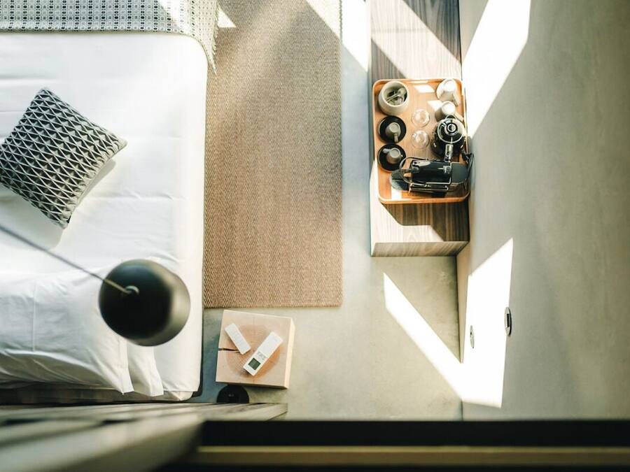 Een net opgemaakt bed met kussens, een nachtkastje met een lamp, een dienblad met kopjes en een camera, en een wandlamp.