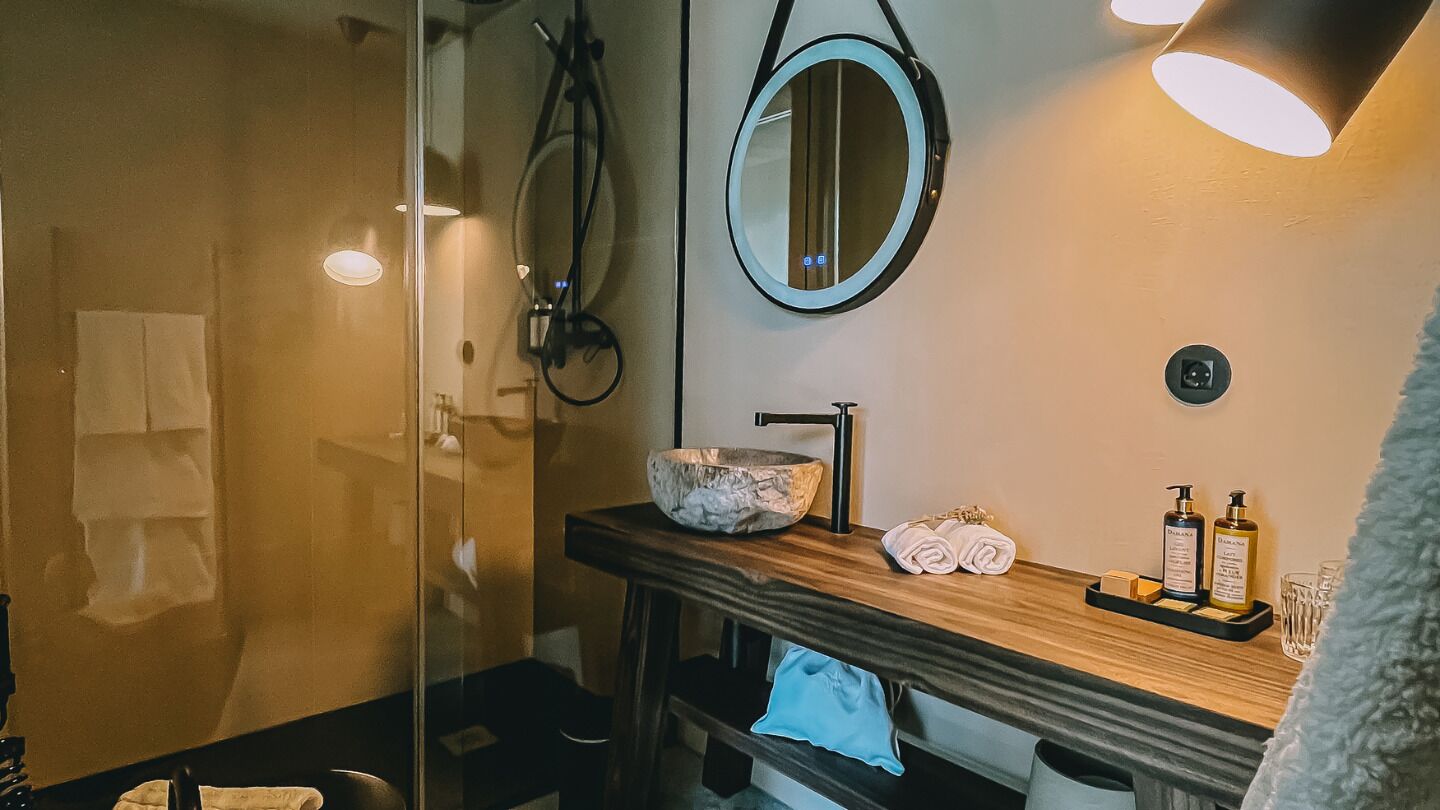 Une salle de bain dotée d’une douche à parois de verre, d’un meuble-lavabo en bois avec un vasque en marbre, d’un miroir rond et de produits de toilette disposés sur le comptoir.