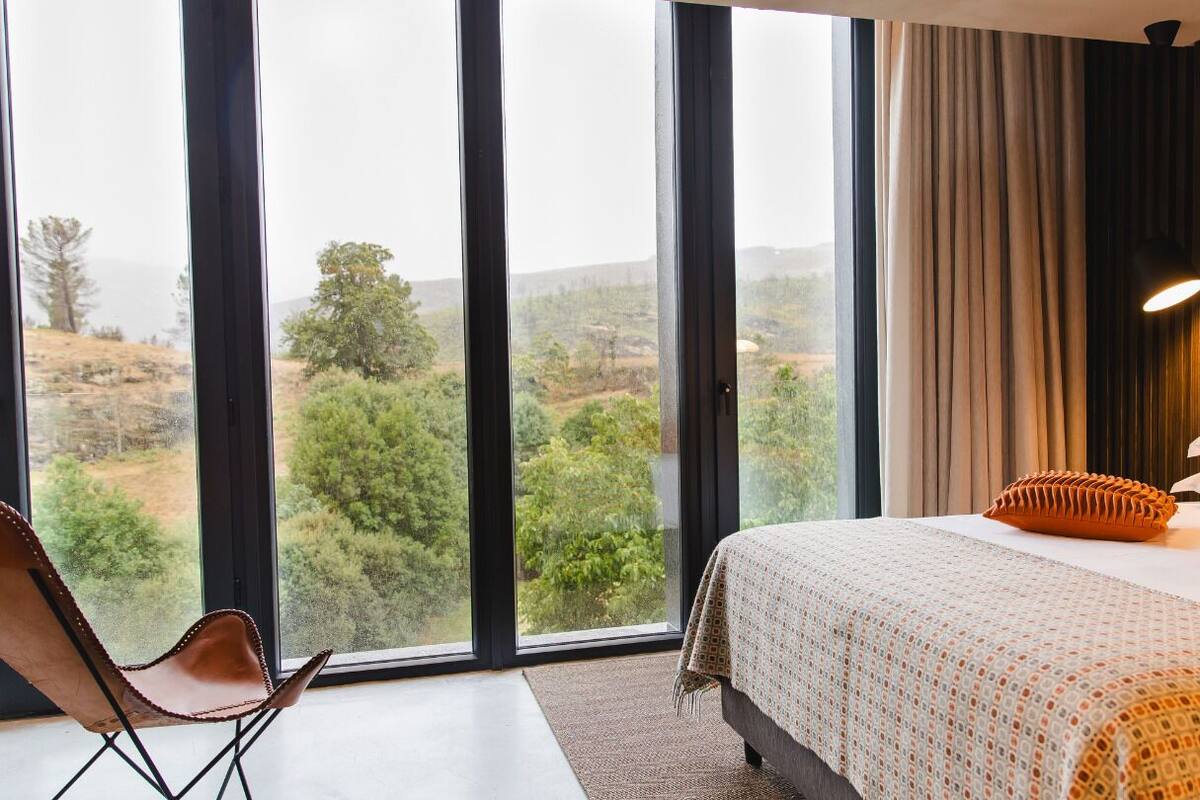 Une chambre avec une grande fenêtre, un lit recouvert d’une literie à motifs et une vue sur un paysage verdoyant.