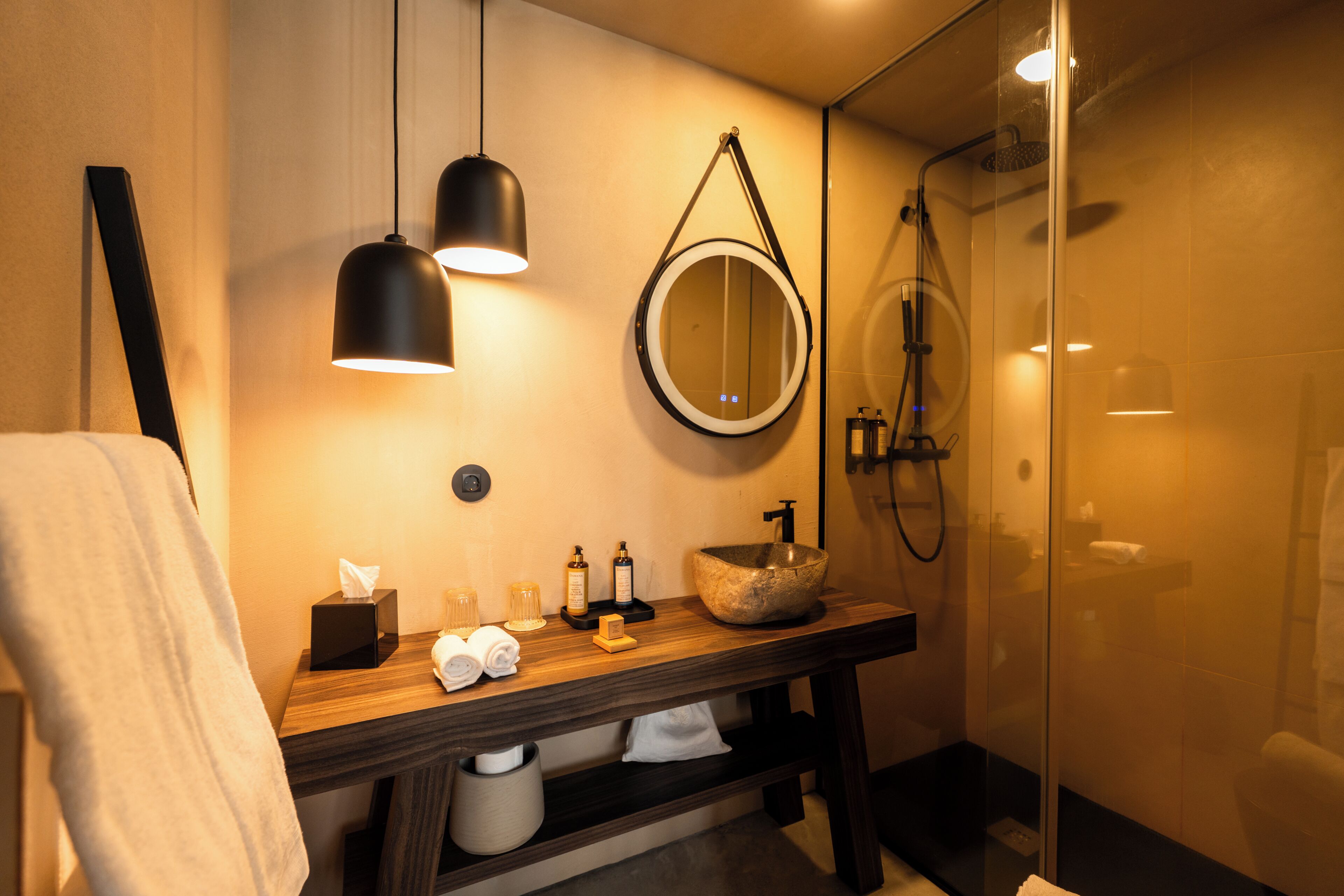 A modern bathroom with a wooden vanity, a round mirror, a stone sink, a handheld showerhead, and a towel rack.