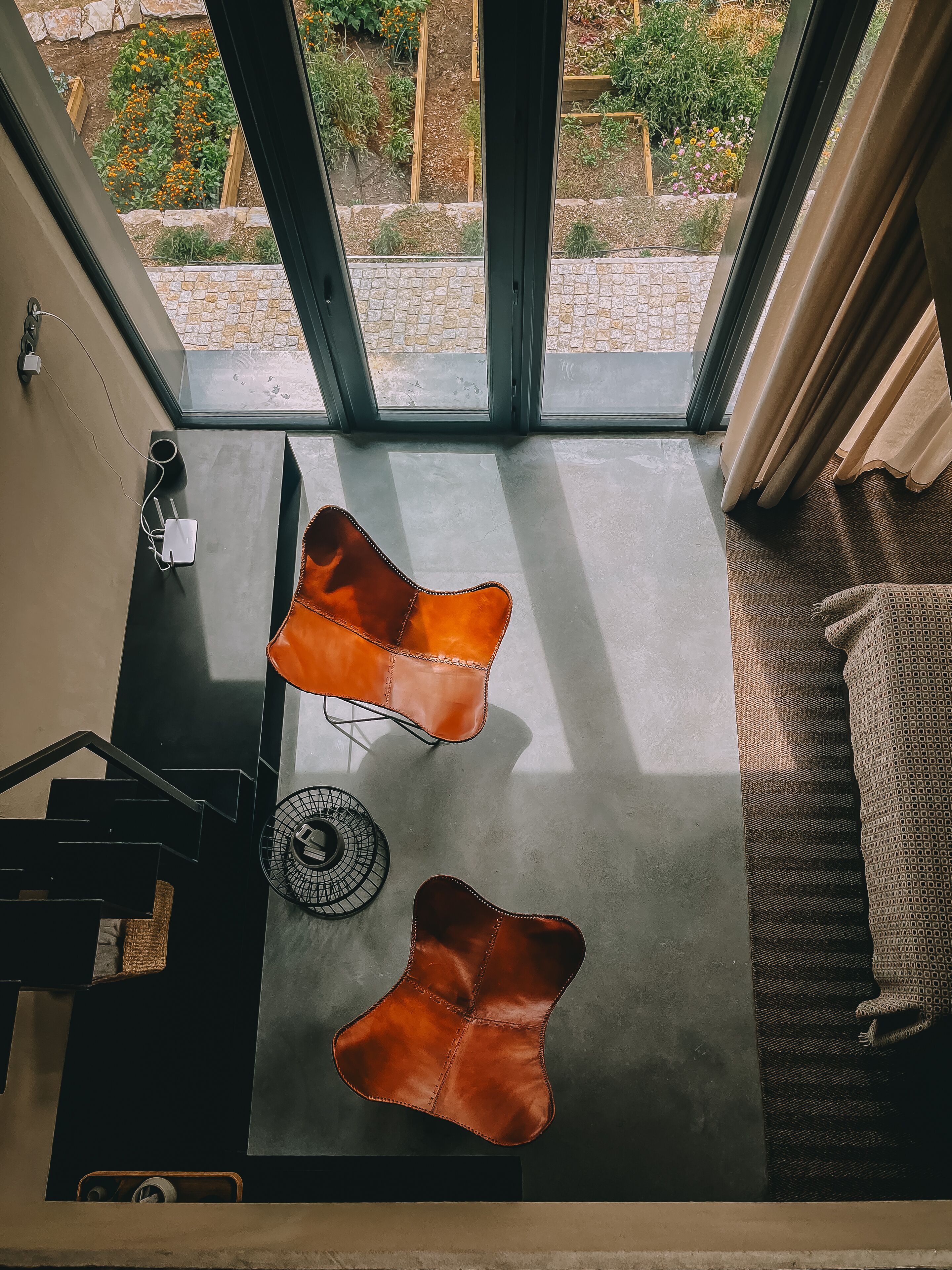 A modern interior with a view of a garden through large windows. There are two leather poufs and a wicker chair.