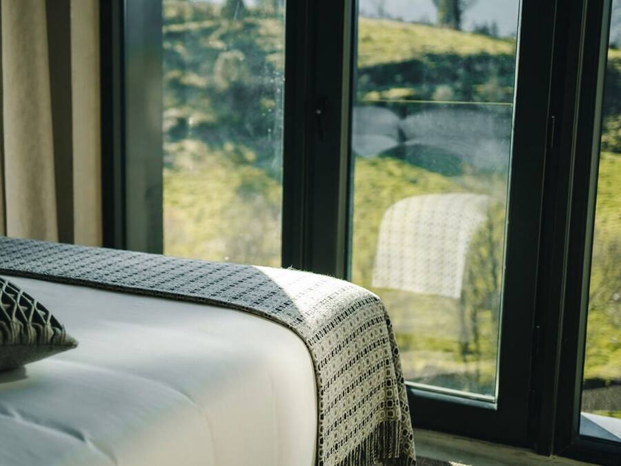 A bed with a white mattress and a gray blanket, positioned next to a window with a view of a green landscape.