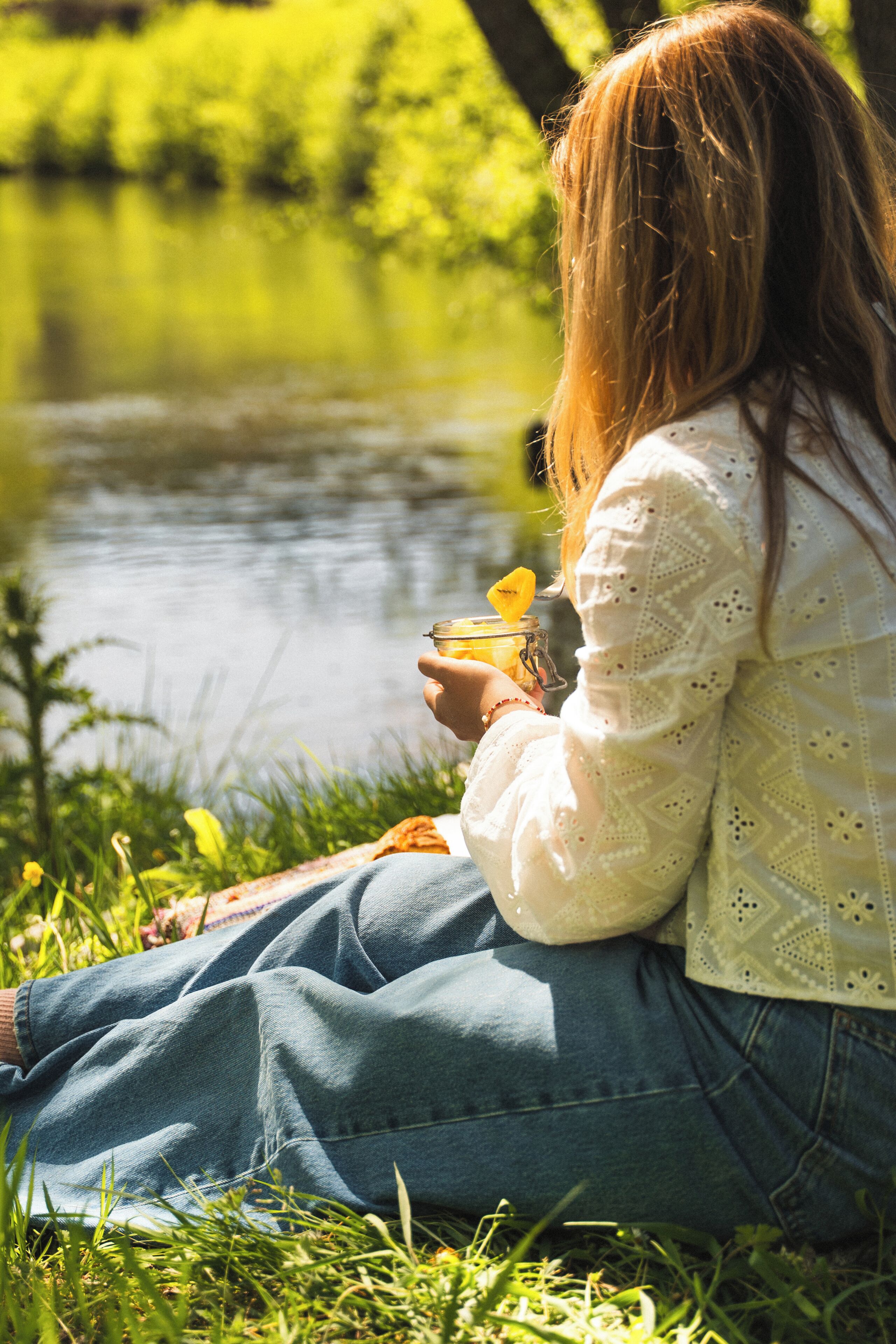 A person sitting by a body of water, holding a jar with a yellow substance.