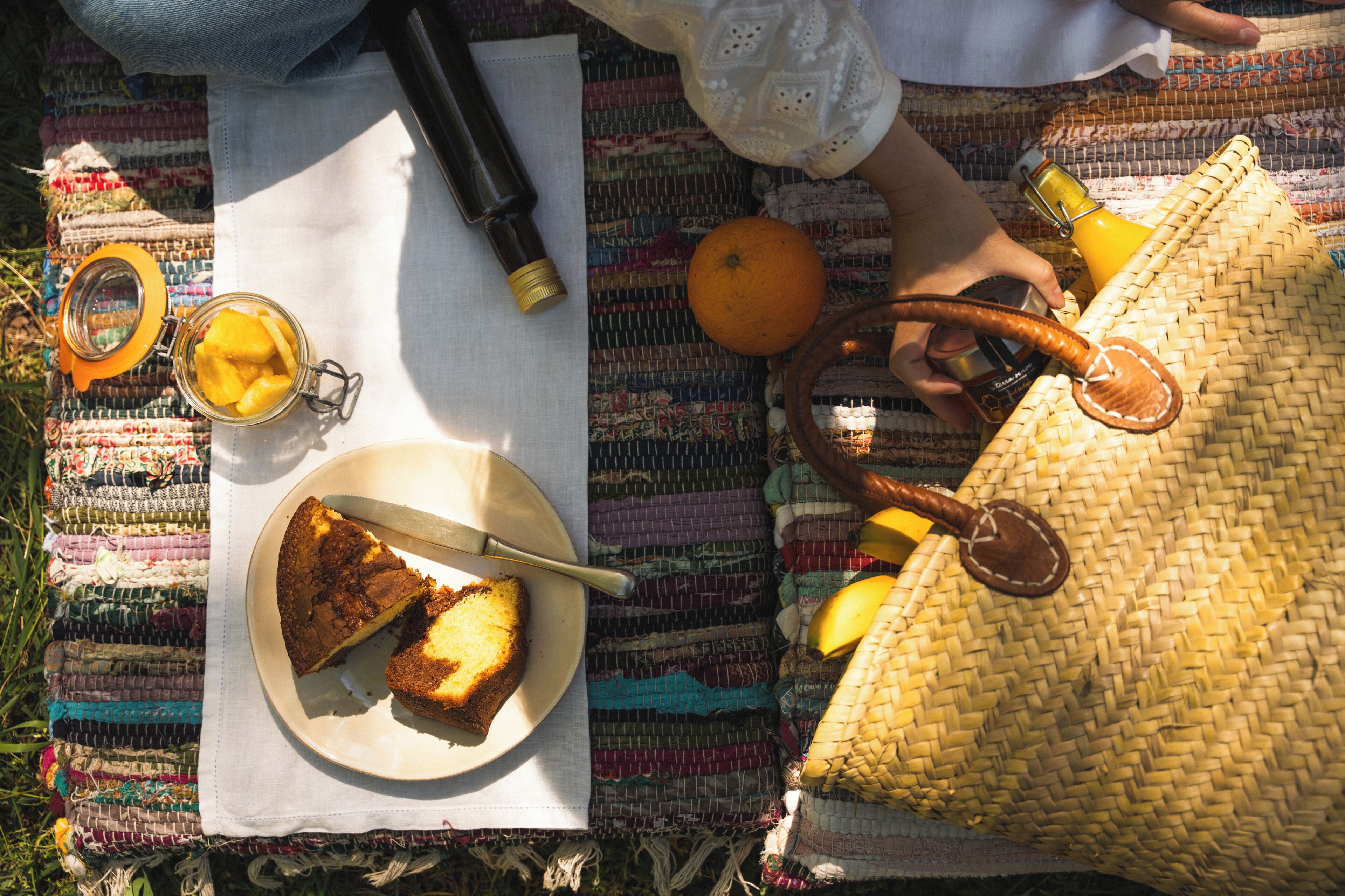 En picnicopsætning med mad på en tallerken, en flaske, et glas og en vævet taske på en farverig måtte.