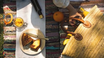 En picnicopsætning med mad på en tallerken, en flaske, et glas og en vævet taske på en farverig måtte.