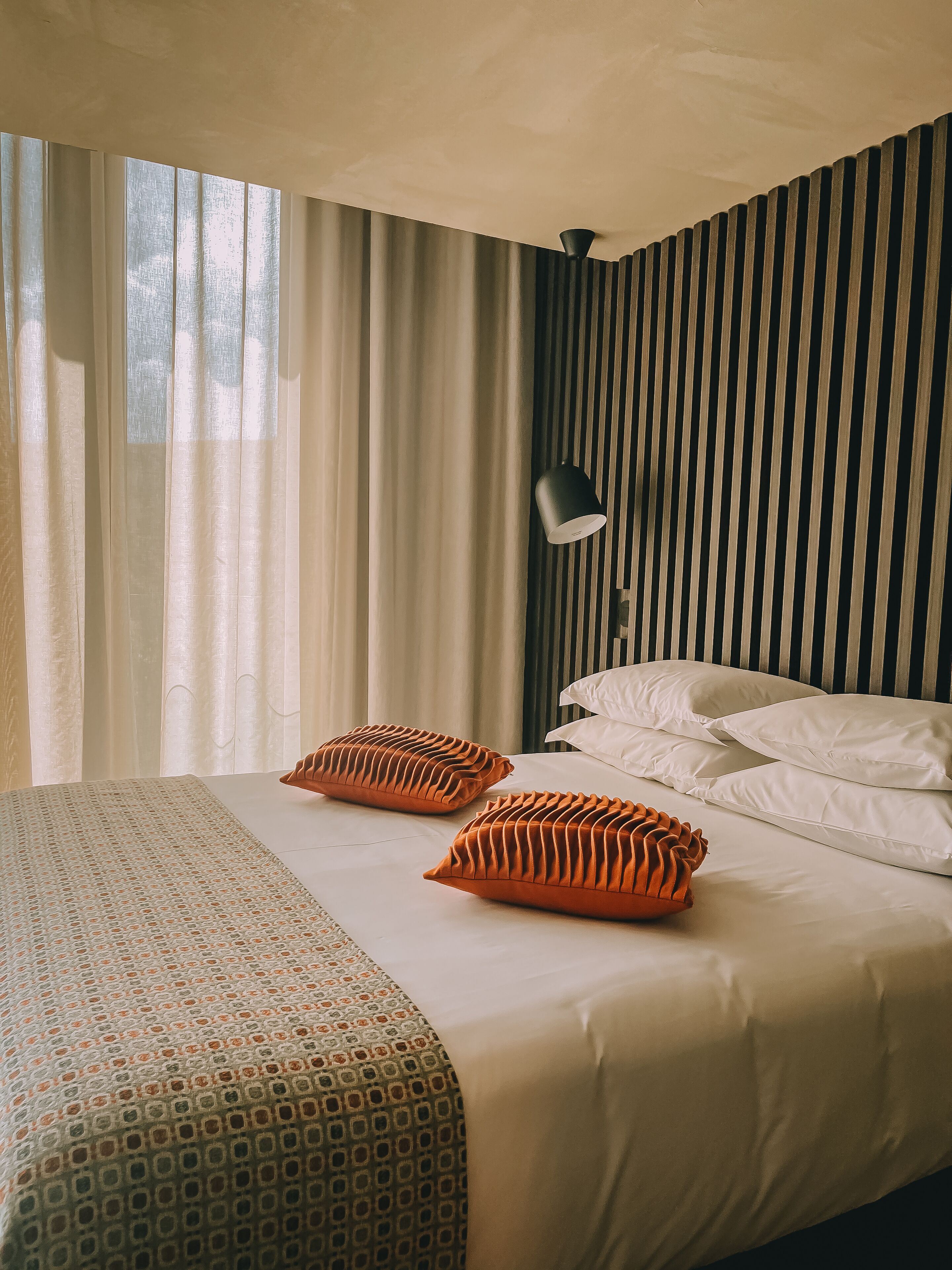 A neatly made bed with patterned bedding and two orange pillows, a bedside lamp, and a window with sheer curtains.
