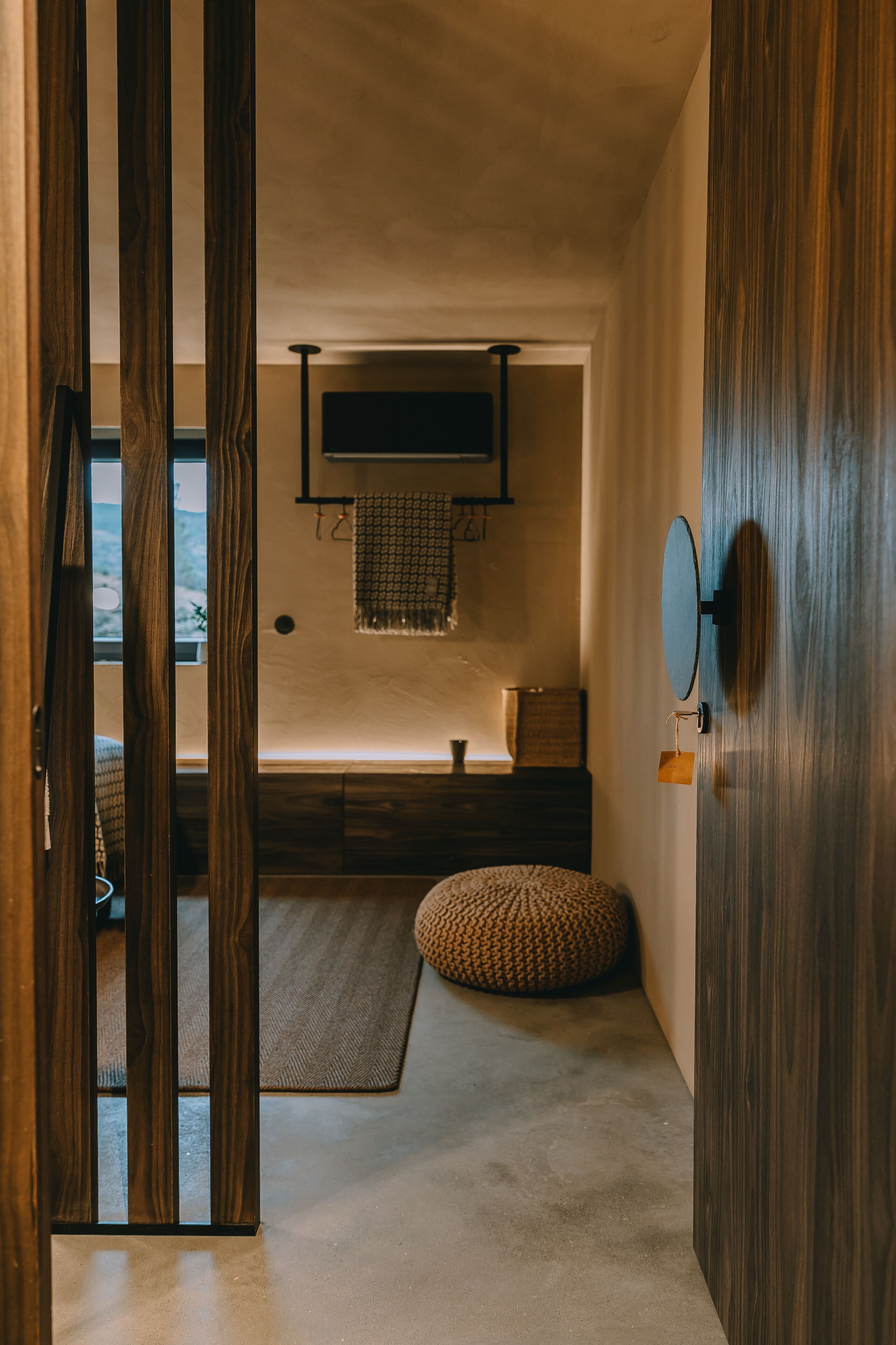 Une pièce avec un plancher en bois, un miroir rond, un petit coussin rond, une étagère fixée au mur et une fenêtre donnant sur un beau paysage.