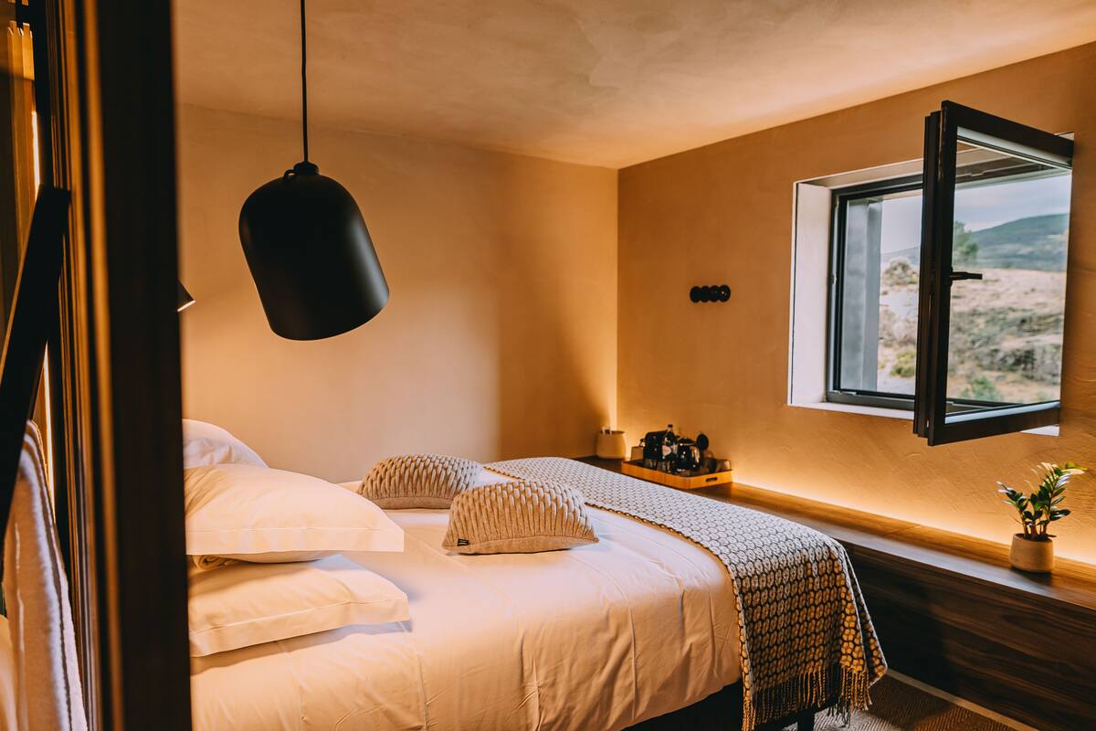 Une chambre à coucher avec un lit, des oreillers, un luminaire suspendu, une fenêtre donnant sur un beau paysage et une petite table avec une plante.