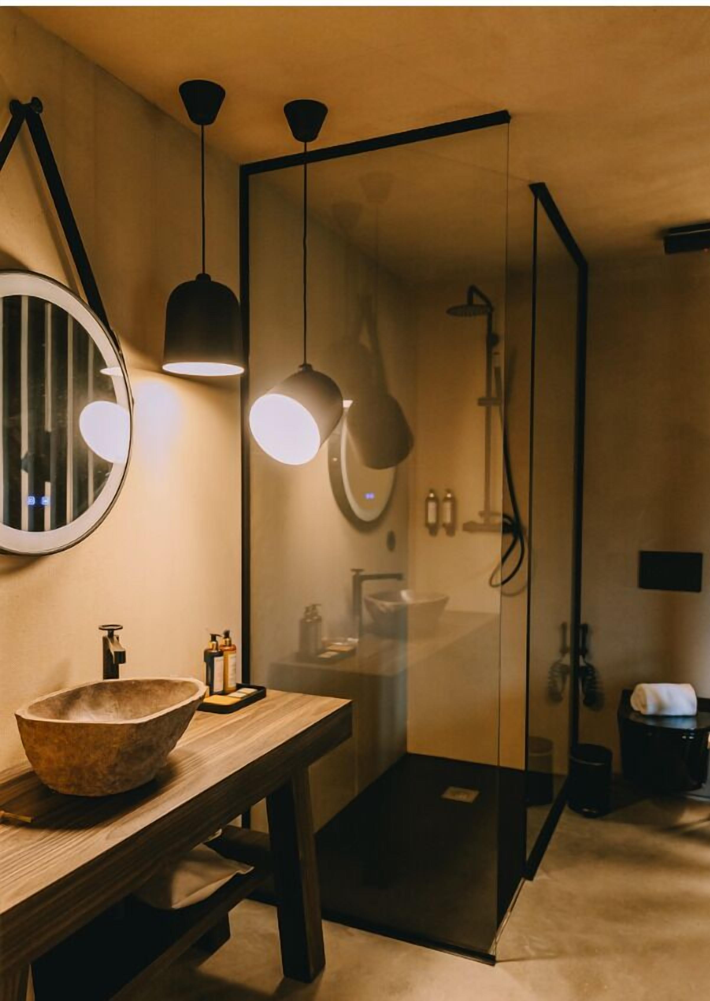 Un baño moderno con tocador de madera, espejo redondo, luces colgantes y zona de ducha con cerramiento de vidrio.