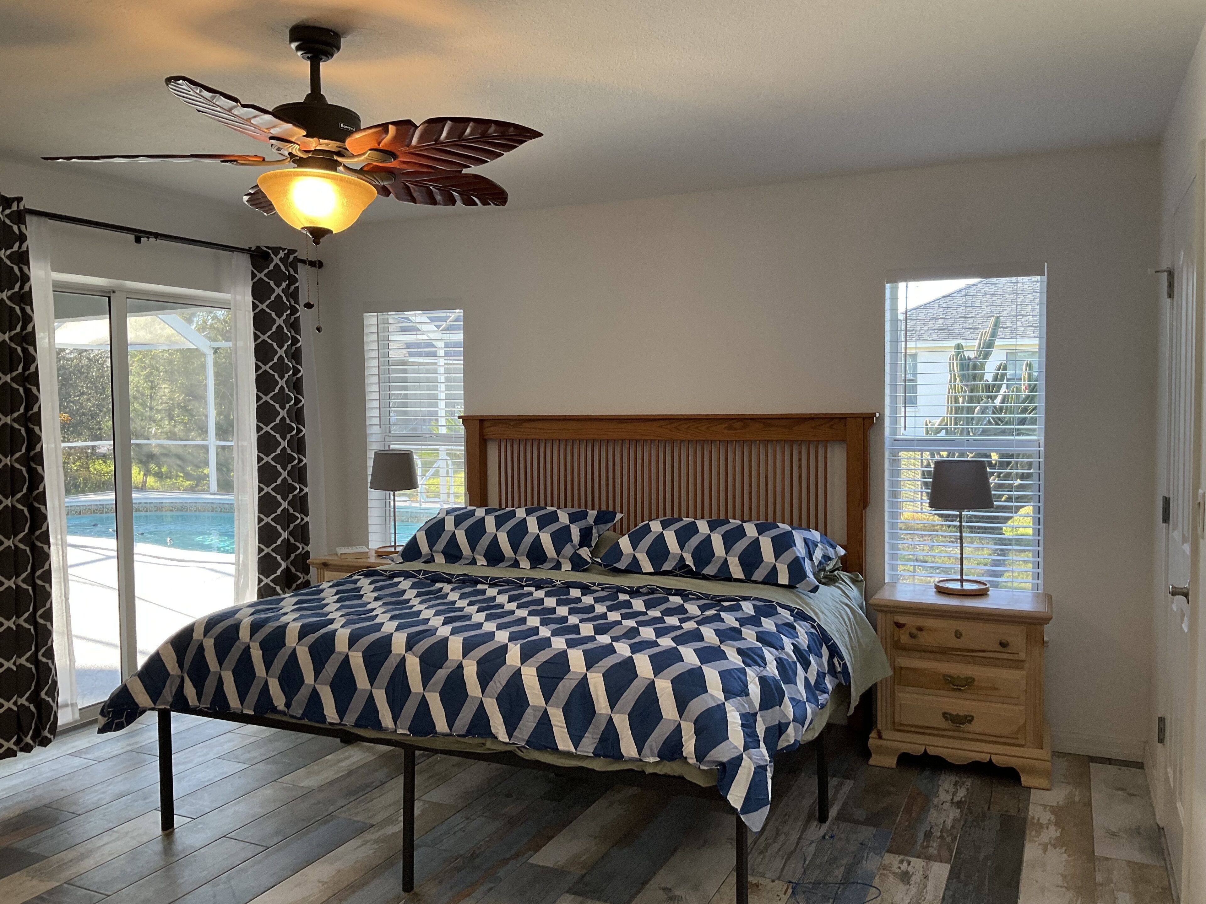 A bedroom with a bed, a ceiling fan, a nightstand, a lamp, and a view of a swimming pool through a window.