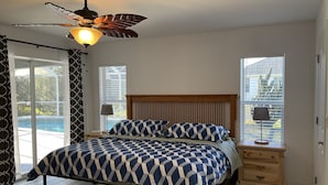 A bedroom with a bed, a ceiling fan, a nightstand, a lamp, and a view of a swimming pool through a window.