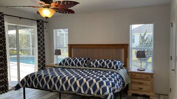 A bedroom with a bed, a ceiling fan, a nightstand, a lamp, and a view of a swimming pool through a window.