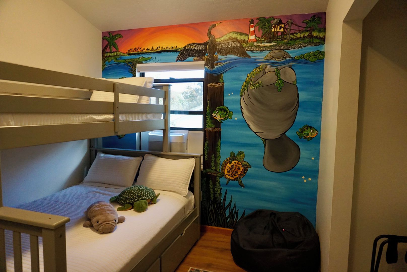 Quarto de beliche infantil com mural de paisagem tropical, um brinquedo de pelúcia em formato de tartaruga e uma bolsa preta no chão.