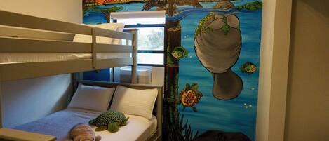Quarto de beliche infantil com mural de paisagem tropical, um brinquedo de pelúcia em formato de tartaruga e uma bolsa preta no chão.
